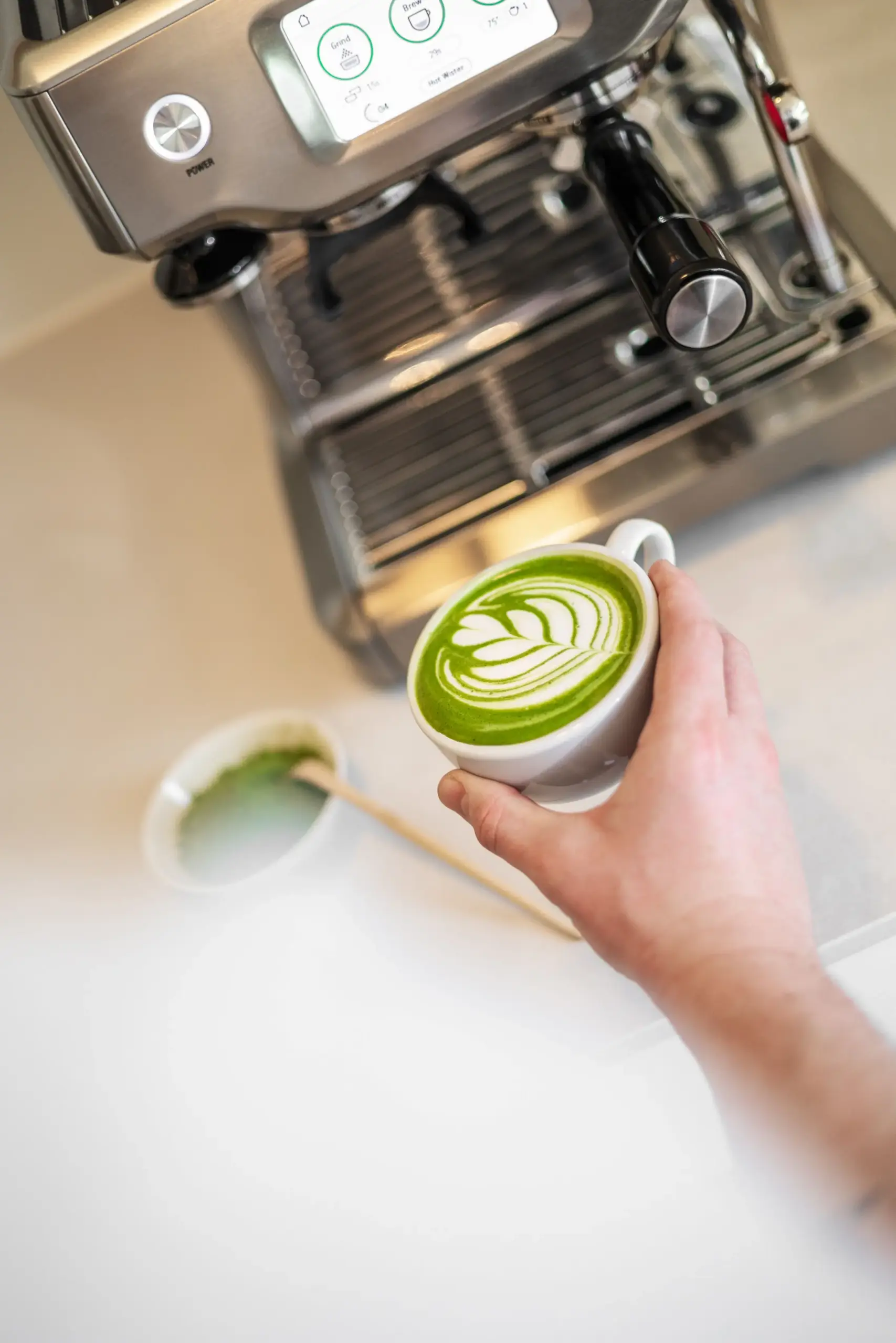 Hand houdt een witte mok met matcha latte art vast, voor een roestvrijstalen espressomachine