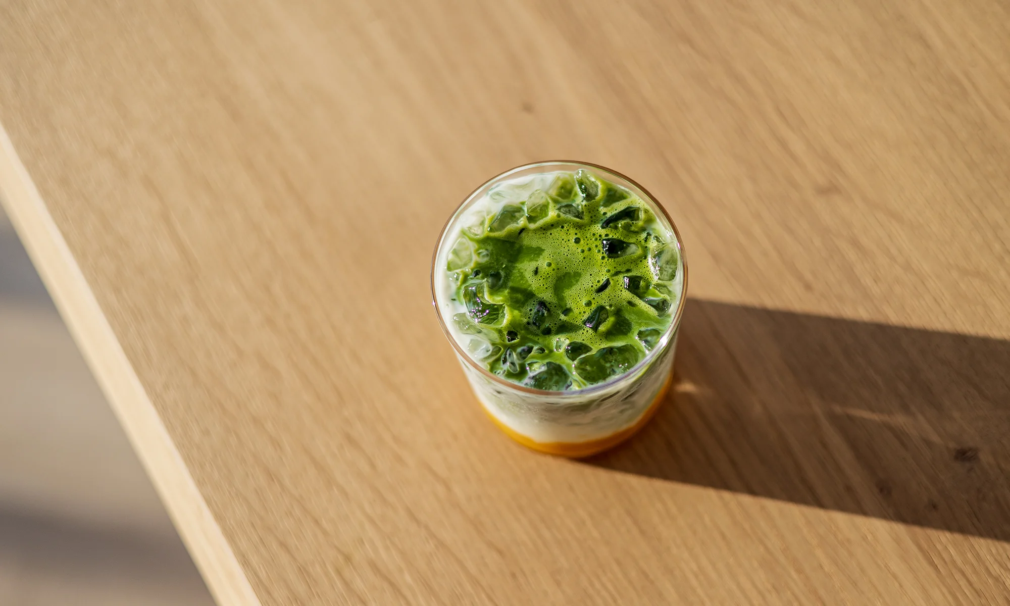 SHINYU Matcha - Bewaarblik 10 Gelaagde ijskoude matcha latte met melk en een oranje siroop op de bodem, gezien van bovenaf op een houten tafel