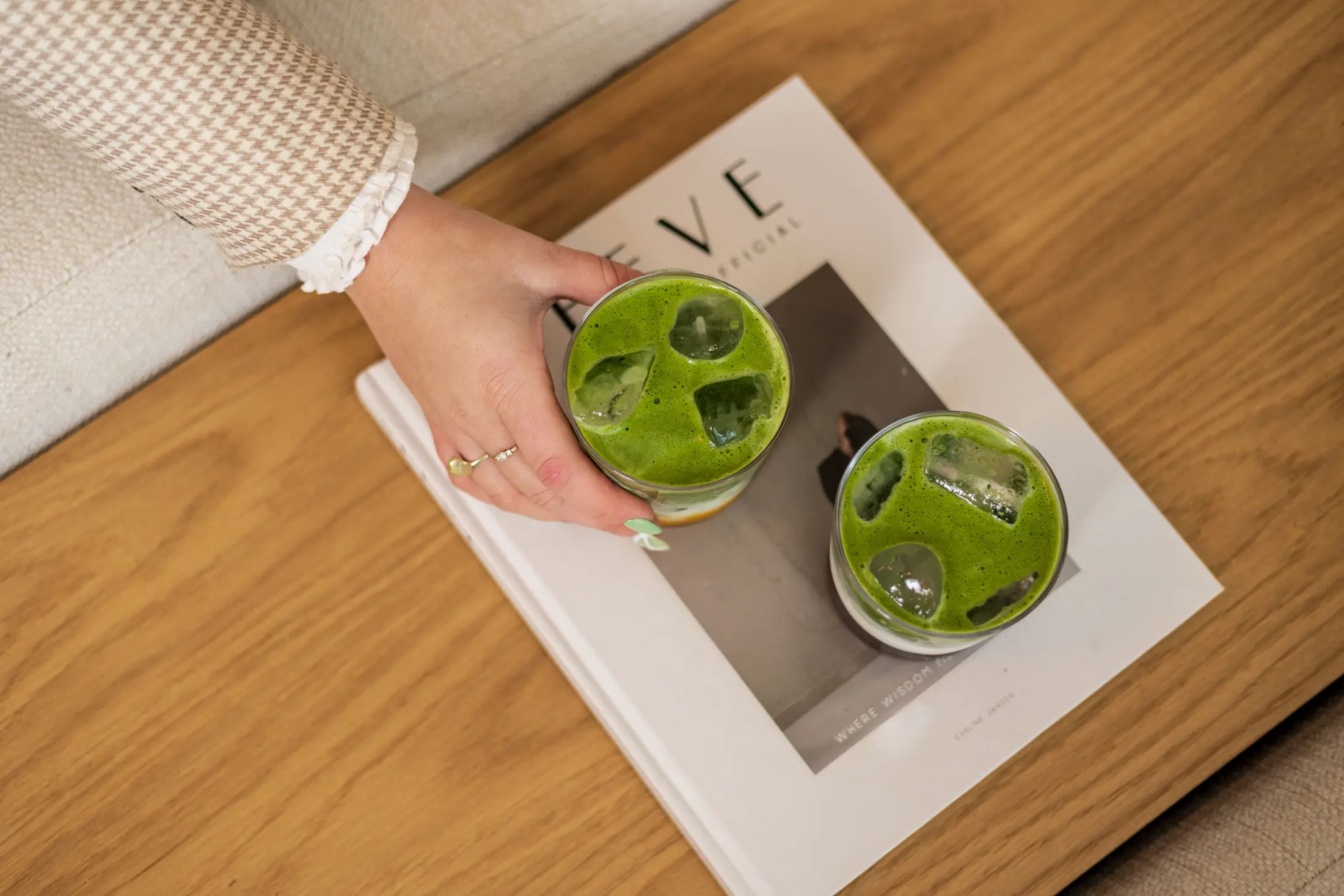 Two iced matcha drinks on a magazine atop a wooden coffee table, with a hand reaching to pick one up
