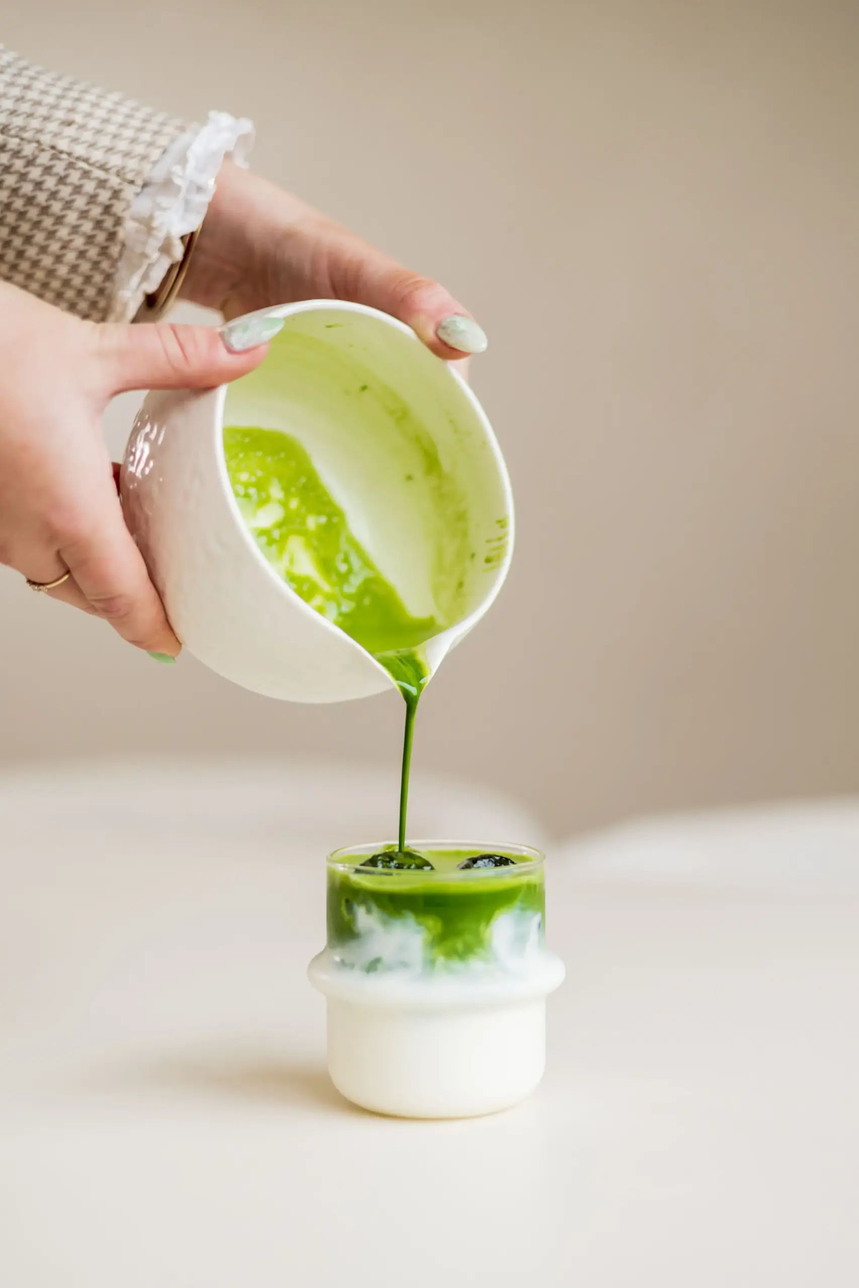Bright green matcha being poured from a ceramic pitcher into a glass over milk, creating distinct layers