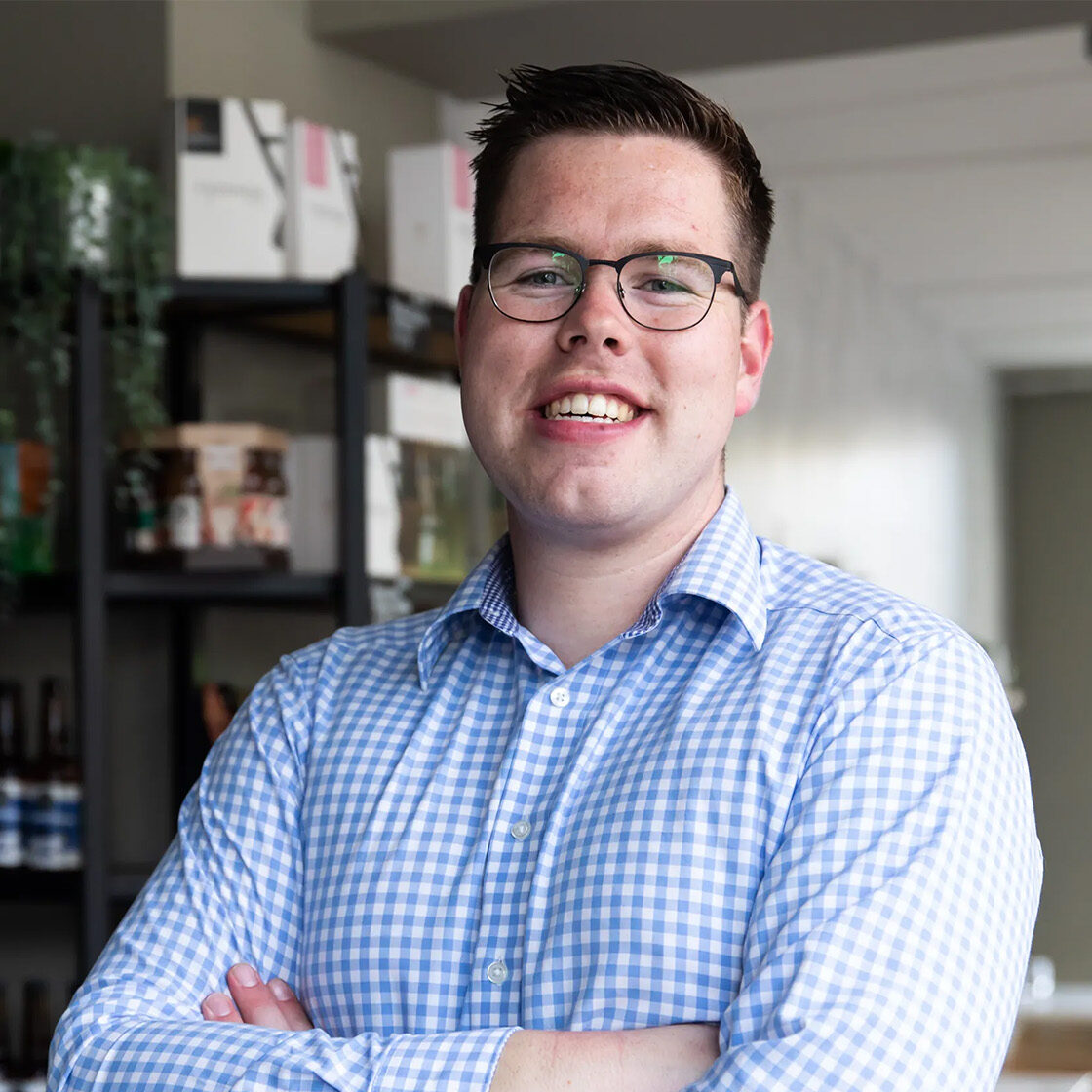 Smiling young man with glasses and arms crossed, wearing a blue gingham shirt, standing in an office or retail space