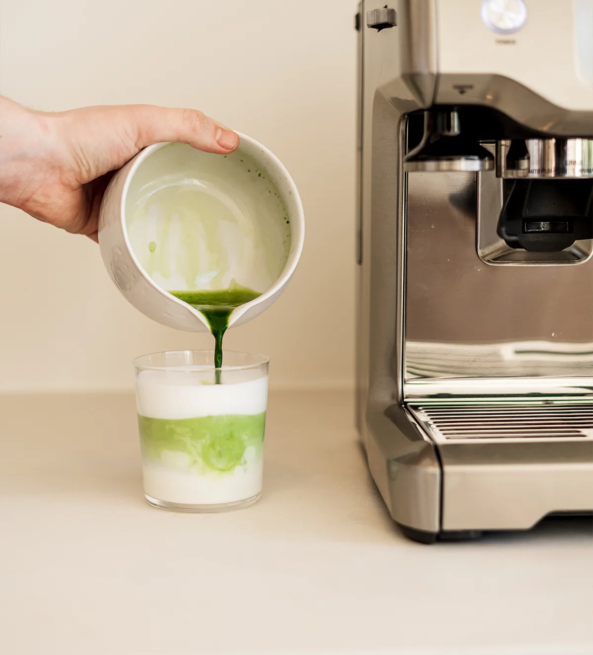Matcha latte bereiden: hand giet matcha over opgeklopte melk in een glas naast een espressomachine
