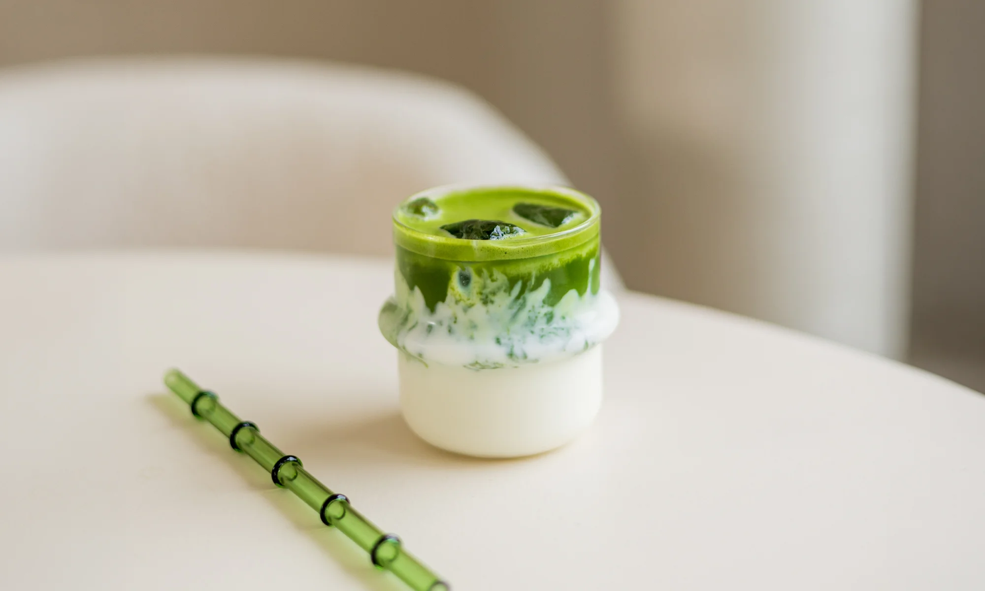 SHINYU Matcha starter kit 5 IJskoude matcha latte met melklagen in een glas, naast een groen glazen rietje op een witte tafel