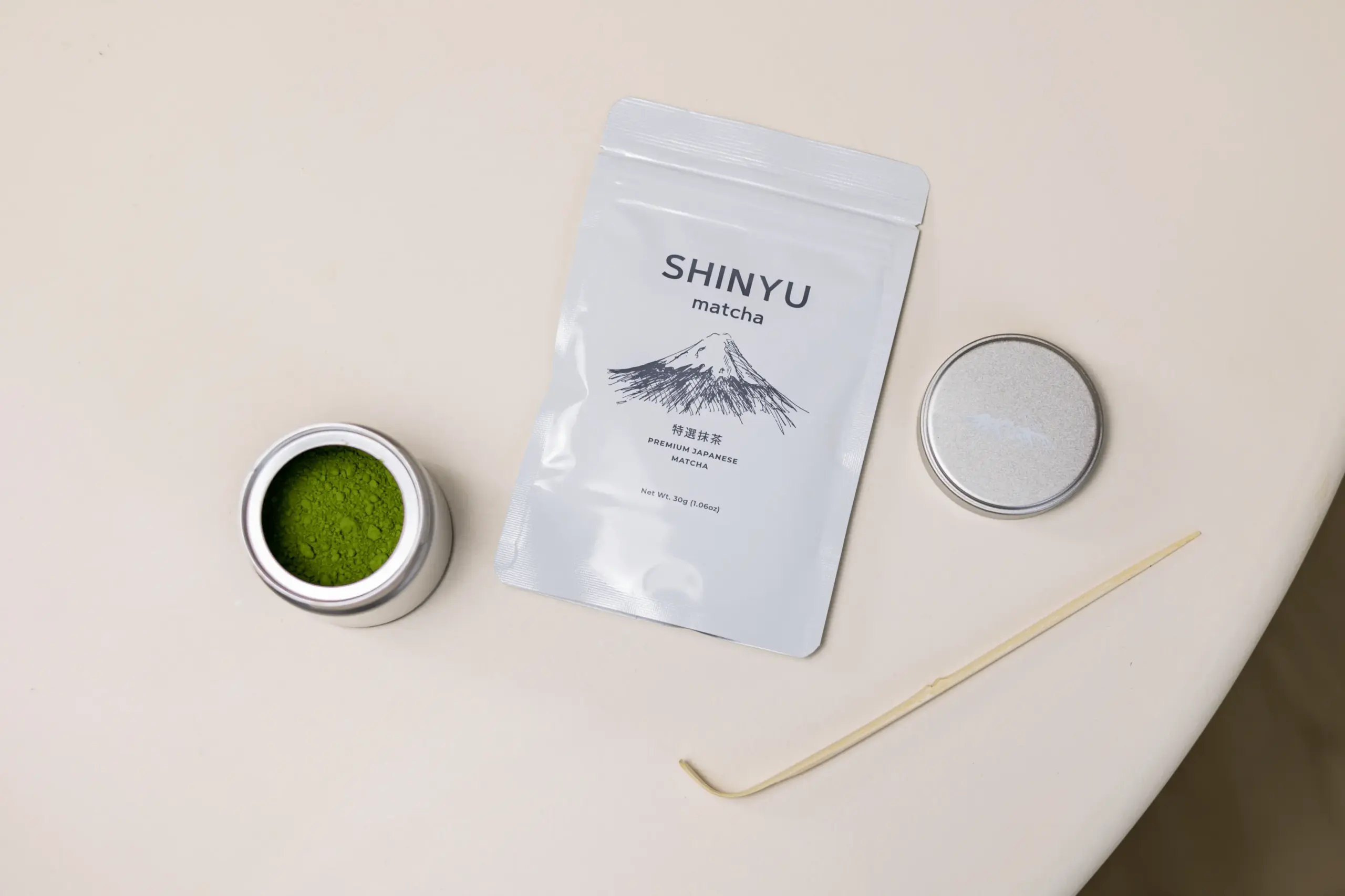 Shinyu Matcha Premium Japanese Matcha zak van 30g, naast een open blikje met groen matchapoeder en een bamboespatel