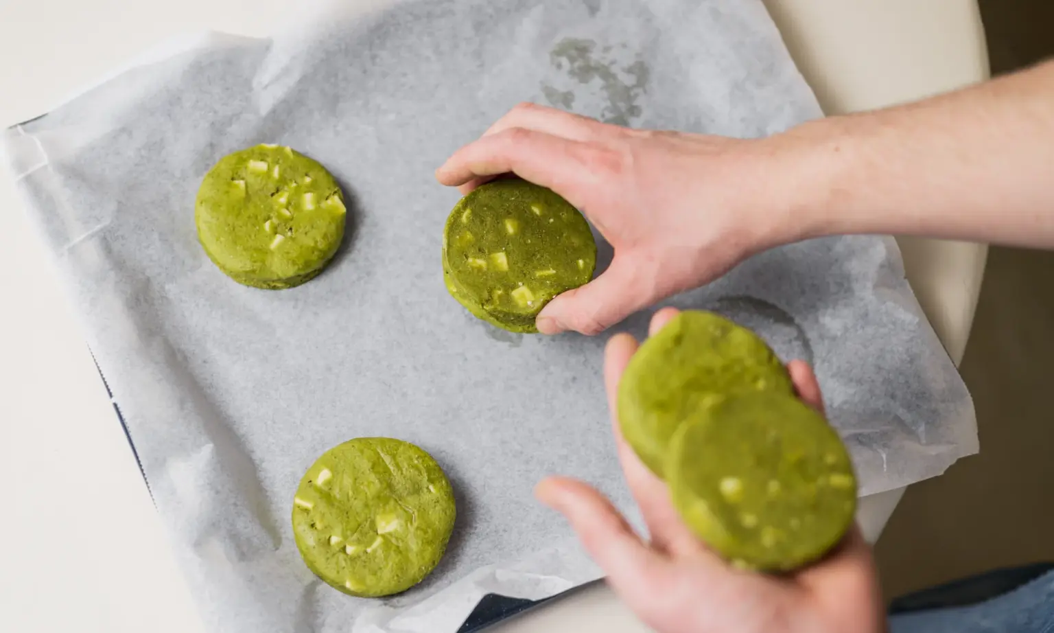 Handen die groene matcha koekjes met witte chocoladestukjes op bakpapier leggen