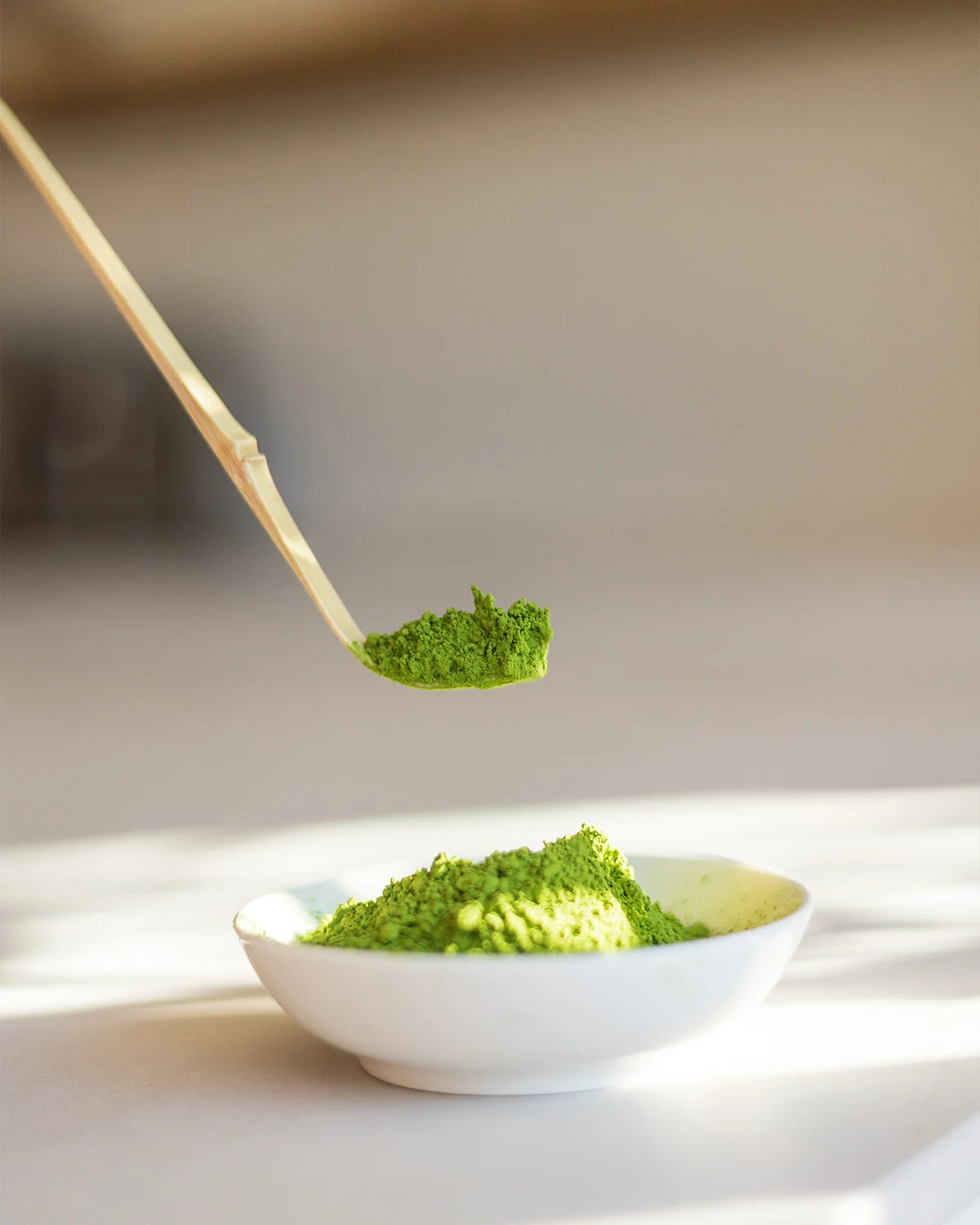 Bamboeschepje met matcha-poeder boven een wit kommetje gevuld met helder groen matcha-poeder