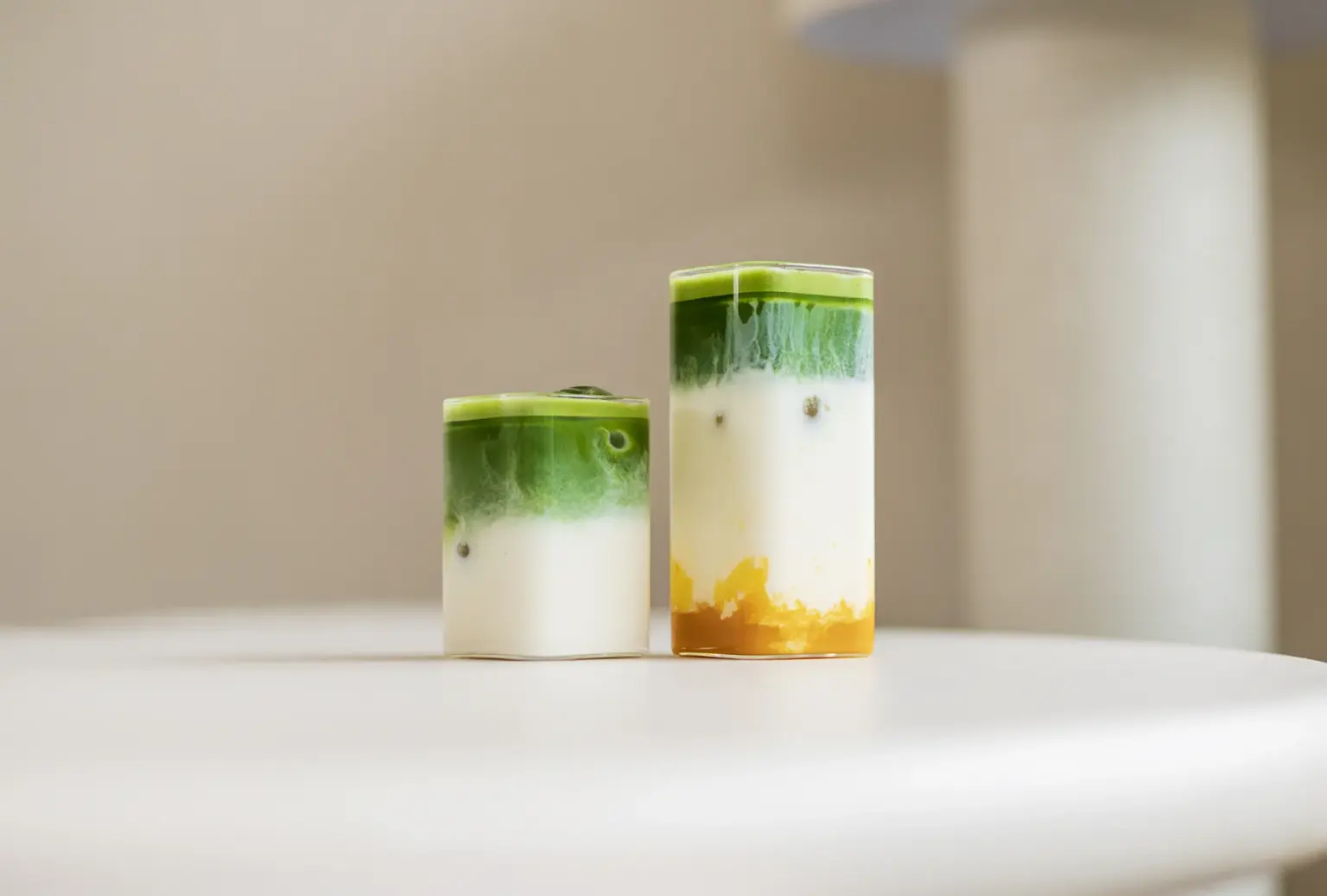 Twee glazen met gelaagde matcha-dranken op een witte tafel, één klein en één groot met een gouden onderlaag