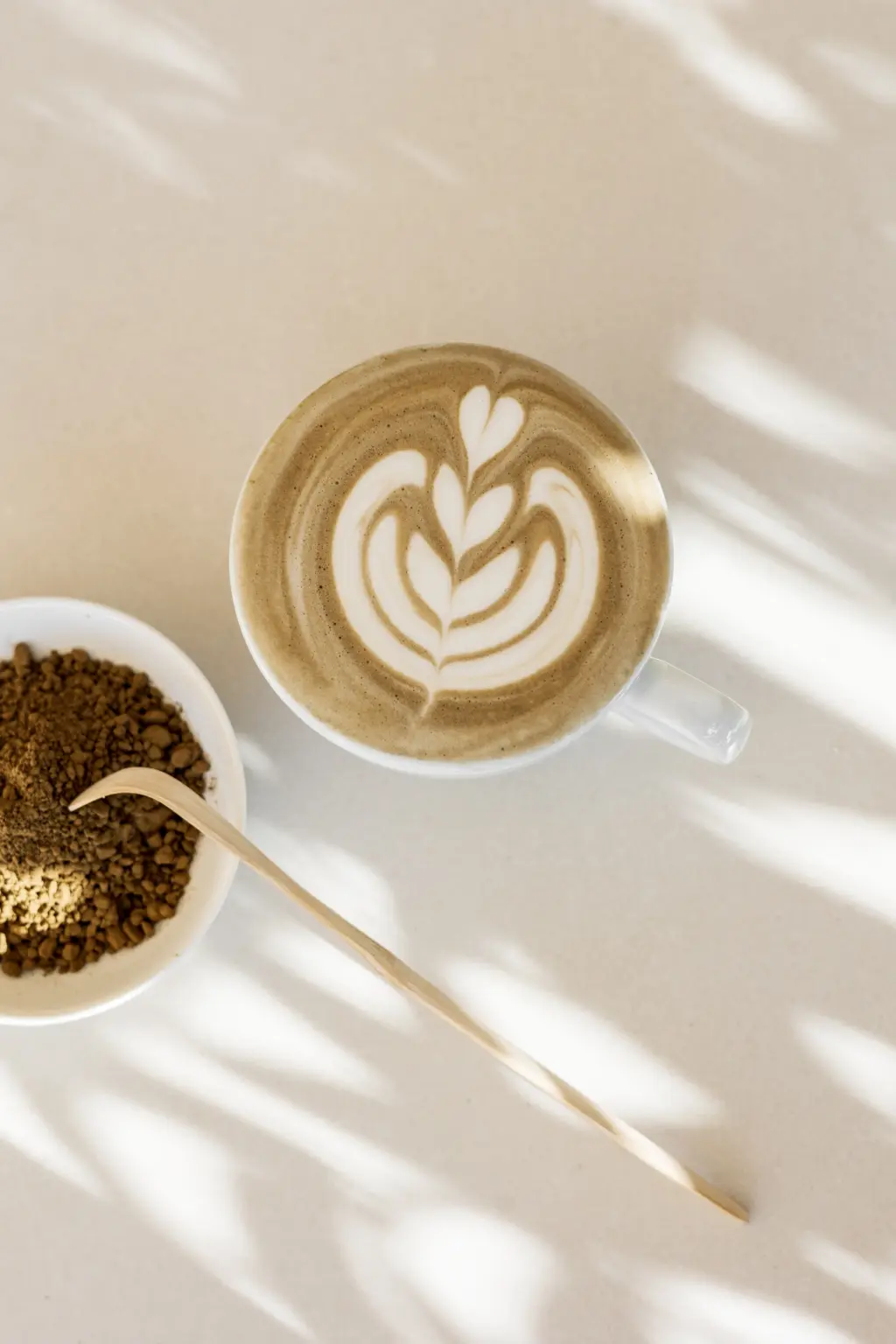 Cappuccino met latte art in een wit kopje, naast een schaaltje koffiekorrels en een houten lepel