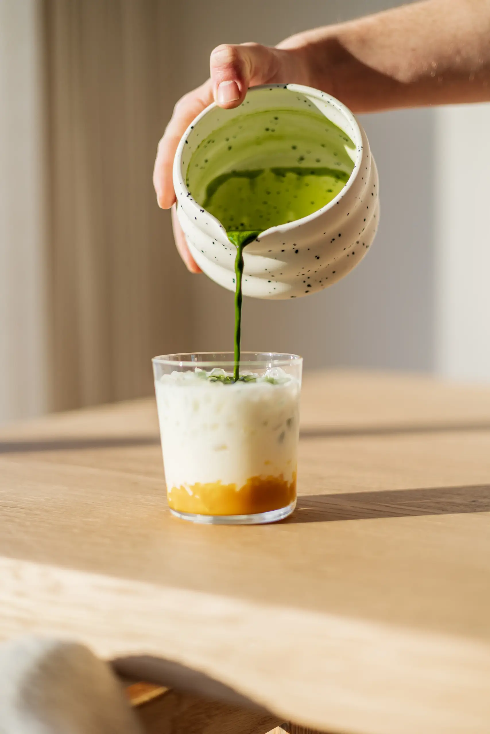 SHINYU Matcha Whisk 4 Matcha wordt uit een keramisch kommetje gegoten in een glas met lagen melk en mangosiroop op een houten tafel