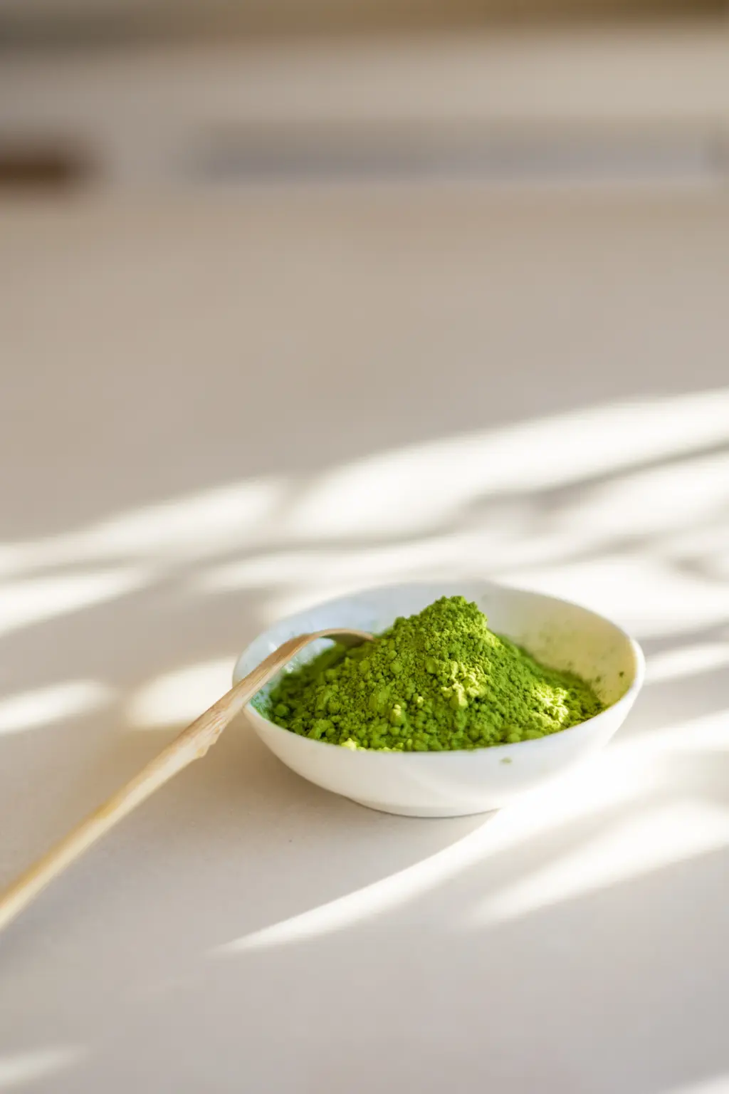 Bright green matcha powder mounded in a small white bowl with a bamboo scoop, set on a sunlit surface