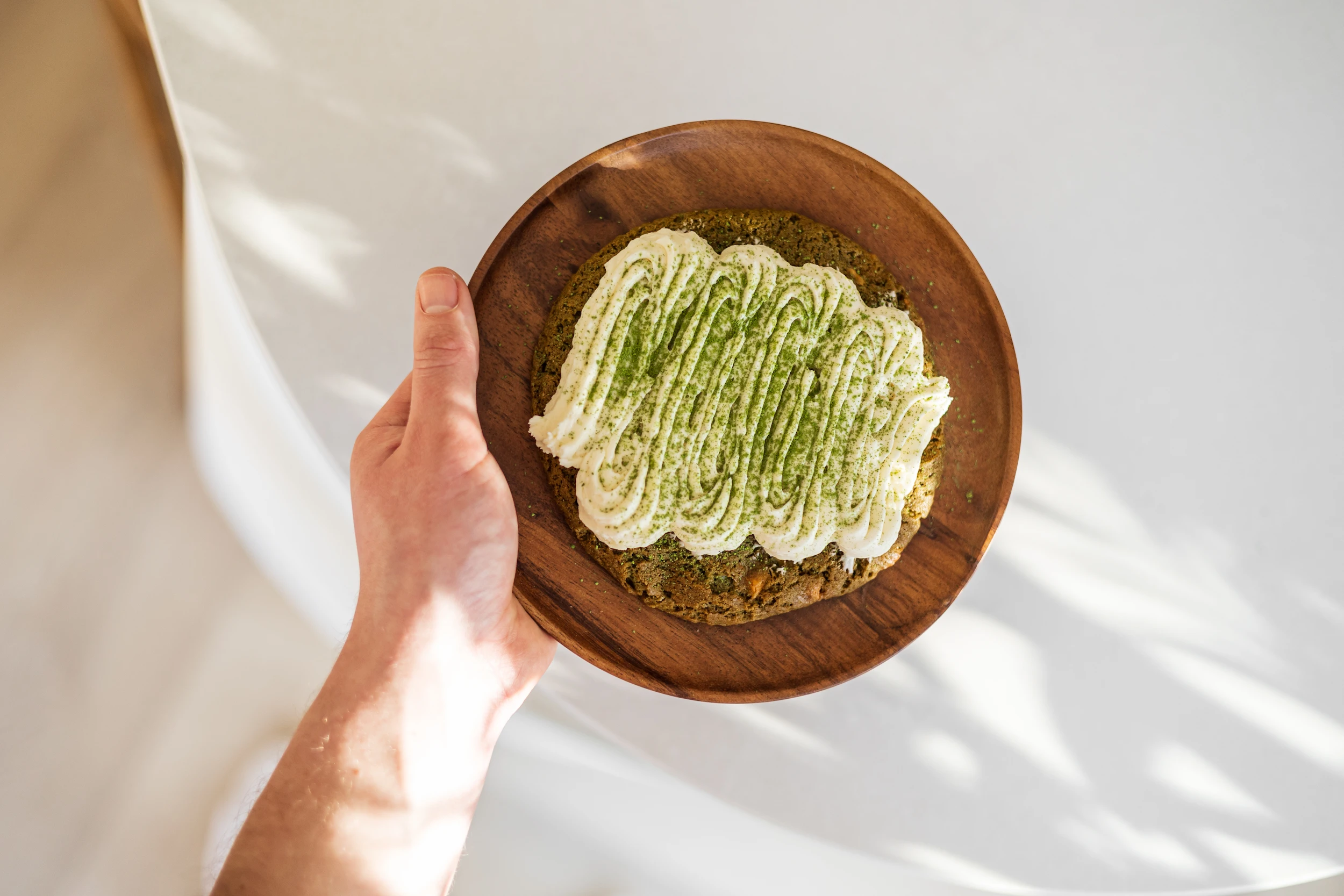 SHINYU Matcha starter kit 7 Hand houdt een houten bord vast met een grote matcha-koek bedekt met witte roomkaasglazuur bestrooid met matchapoeder