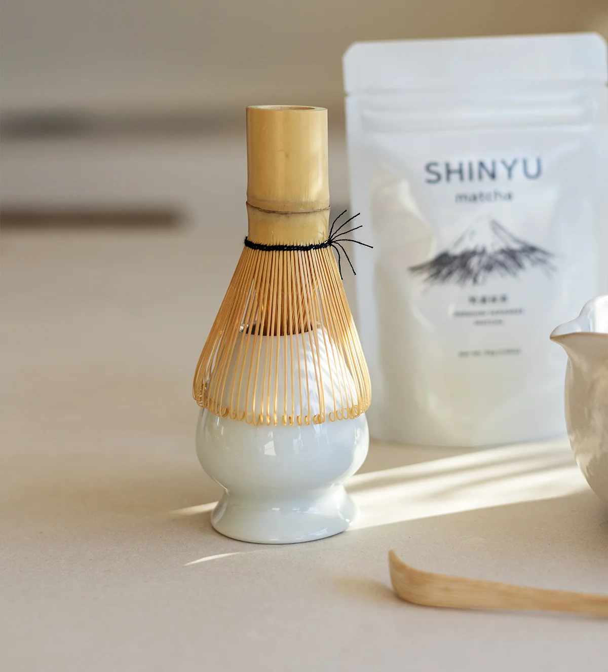 SHINYU Matcha starter kit 8 Bamboe matcha-klopper op een wit keramisch standaard, met een zak Shinyu Matcha en een bamboeschepje op de achtergrond