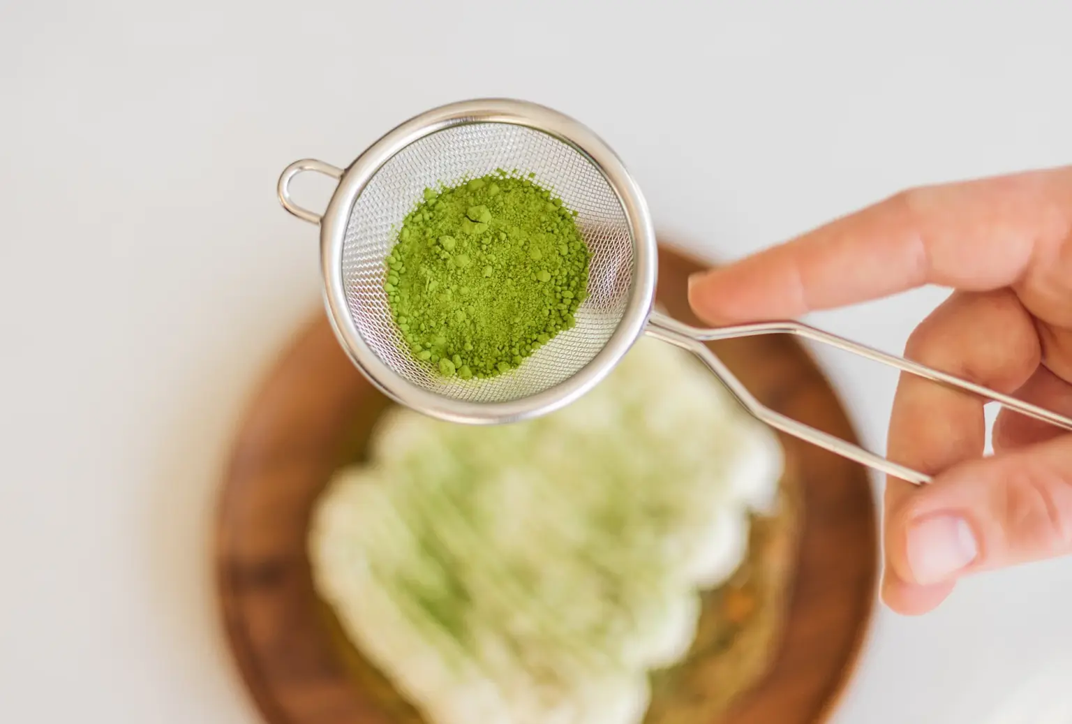 Hand die een fijnmazige zeef met helder groen matchapoeder vasthoudt boven een houten schaal met een Japans gebakje