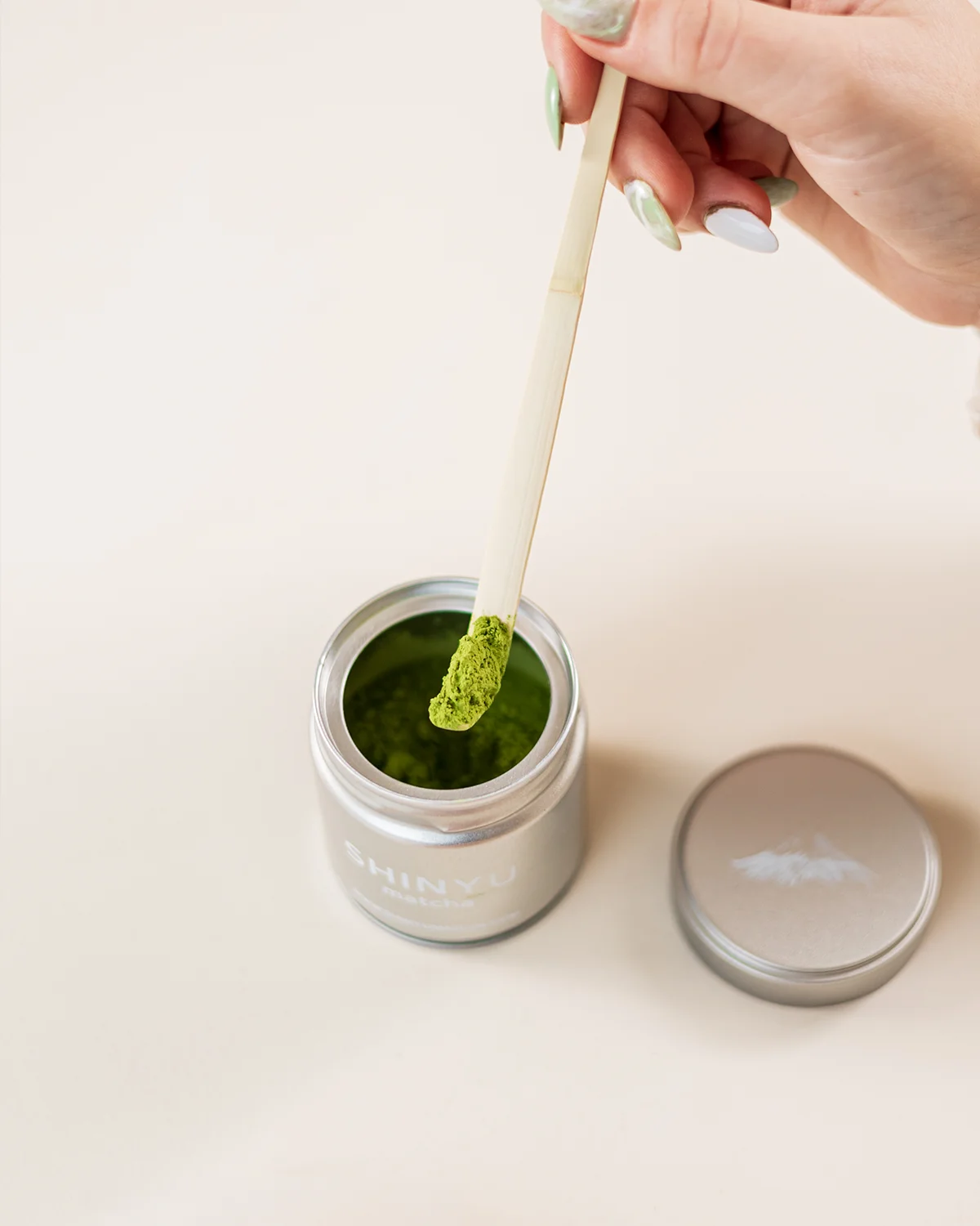 Hand die een bamboeschepje met matcha-poeder uit een zilveren Shinyo-blik haalt