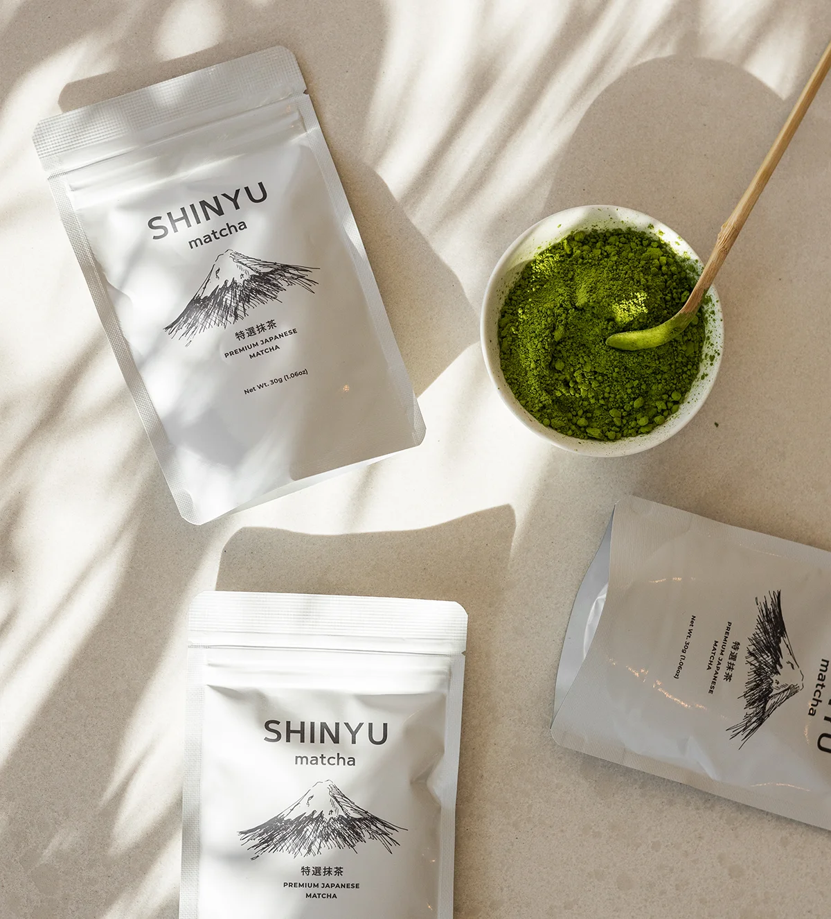 Three Shinyu Matcha Premium Japanese Matcha pouches arranged on a neutral surface beside a small bowl of vibrant green matcha