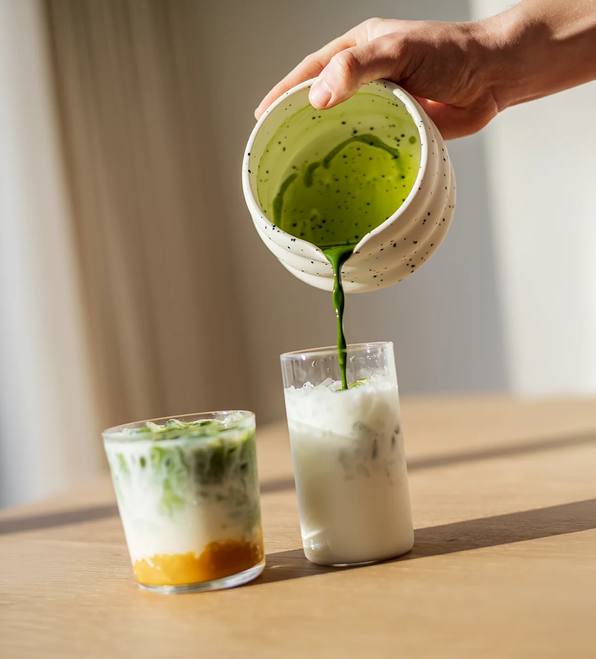 SHINYU Matcha - Bewaarblik 9 Matcha wordt vanuit een keramisch kommetje gegoten over een glas met melk en ijs, naast een tweede glas met matcha-latte