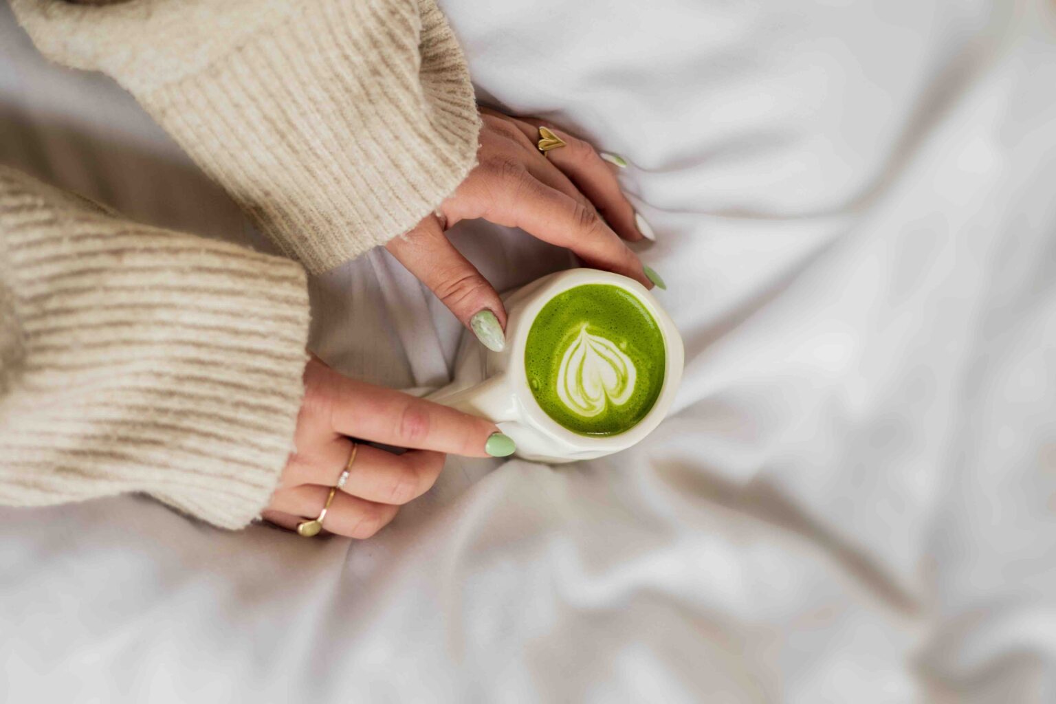 Handen in een beige trui houden een kop matchalatte met een hartvormig melkpatroon vast op een wit bed
